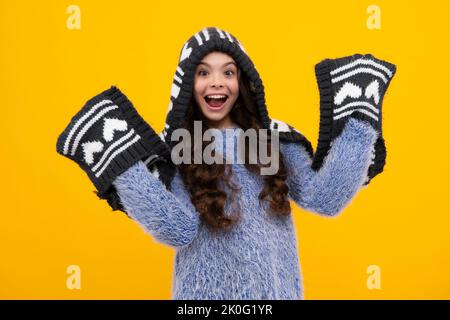Amazed teenager. Warm hat with hood and scarf. Modern teen girl wearing sweater and knitted hat on isolated yellow background. Excited teen girl. Stock Photo