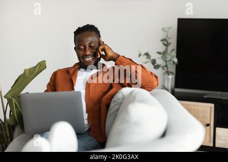 Home office concept. Happy black man working on laptop while sitting on comfy sofa at home interior, copy space Stock Photo