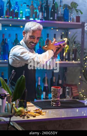 Professional bartender in a white shirt and black apron holds a steel shaker in his hands. Blurred bottles on shelves in background Stock Photo