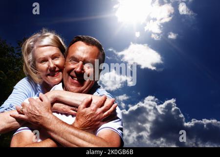 Senior woman hugging husband from behind against cloudy sky. Portrait of a pretty woman hugging husband from behind against cloudy sky. Stock Photo