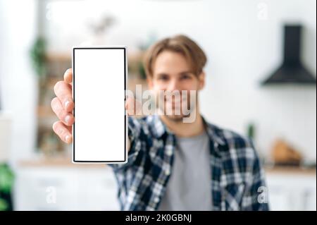 Defocused positive stylish modern caucasian young man, in casual clothes, stands in the living room on the background of the kitchen, shows a smartphone with a blank white mock-up screen, smiles Stock Photo