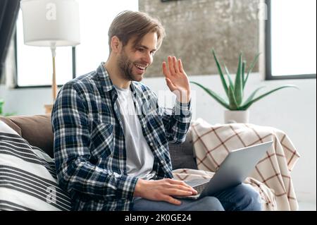 Handsome caucasian man sits on a couch talks online. Attractive freelancer guy using laptop, have a video call, communicate with colleagues or friends by video conference, waving hand and smiling Stock Photo