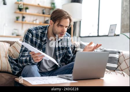 Puzzled confused caucasian young man in stylish casual clothes, freelancer, web designer, sitting in living room on the sofa, working on a laptop on a project, analyze documents, misunderstanding Stock Photo
