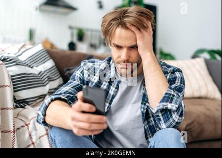 Frustrated unhappy caucasian young stylish man, sitting on a floor near a sofa in living room, sadly looking at smartphone screen, feel loneliness, got bad news or message, despair emotions Stock Photo