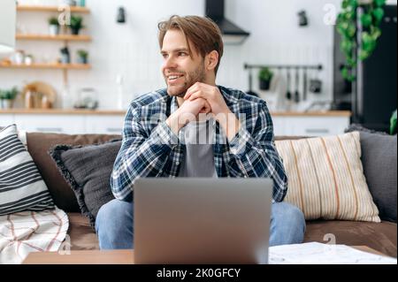 Satisfied handsome young caucasian man in casual stylish clothes, sitting on the couch at home in cozy living room, using a laptop, working or study online, looking to the side, dreaming, smiling Stock Photo