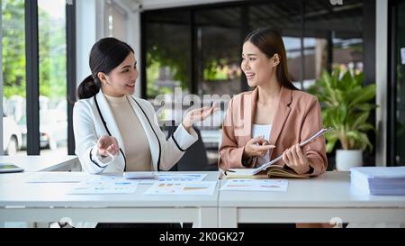 Two cheerful and attractive young Asian businesswomen having a good conversation while working together in the office. Stock Photo