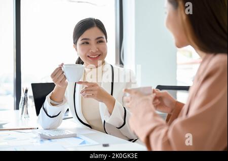 Gorgeous young Asian female boss or marketing manager having a good conversation with her assistant during the coffee break in the office. Stock Photo
