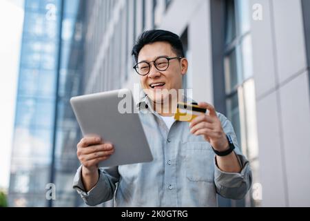 Mobile shopping. Excited korean businessman using tablet and credit card, making payment against office building Stock Photo