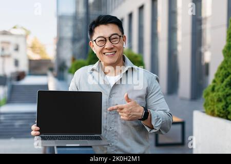 Portrait of korean businessman pointing at laptop computer with blank black screen, standing outdoors, mockup Stock Photo