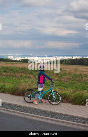 A little girl on her bike and stopped looks at thecityscape in front of her. The clouds are gray blue and threatening. She wears an helmet Stock Photo