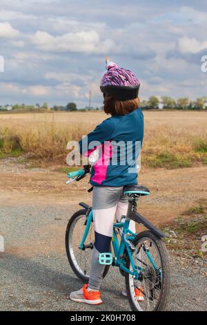 A stopped little girl on her bike points to the threatening sky. The clouds are gray blue. The helmeted child is from behind. Stock Photo