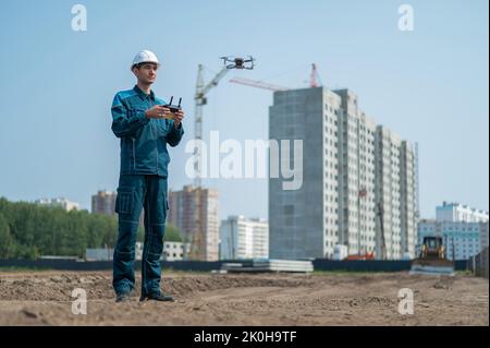 A man in a helmet and overalls controls a drone at a construction site. The builder carries out technical oversight. Stock Photo