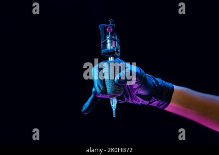 Closeup of tattooer master's hand in black glove holding machine for making tattoo art on body isolated on dark background in neon light. Stock Photo