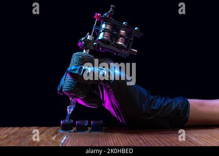 Closeup of tattooer master's hand in black glove holding machine for making tattoo art on body isolated on dark background in neon light. Stock Photo