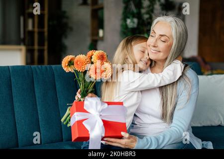 Glad caucasian little granddaughter hugging aged grandma with bouquet of flowers and box gift Stock Photo