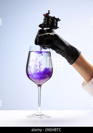 Wine glass and purple ink. Tattooer master's hand in black glove holding machine for making tattoo art on body isolated on white background Stock Photo
