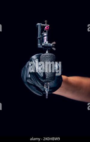 Machine for tattoo. Human hand in black protective glove holding professional equipment for making tattoo on body isolated on dark background. Stock Photo