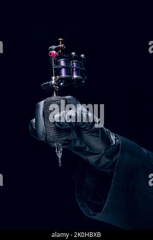 Machine for tattoo. Human hand in black protective glove holding professional equipment for making tattoo on body isolated on dark background. Stock Photo