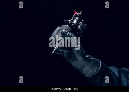Machine for tattoo. Human hand in black protective glove holding professional equipment for making tattoo on body isolated on dark background. Stock Photo
