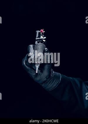 Machine for tattoo. Human hand in black protective glove holding professional equipment for making tattoo on body isolated on dark background. Stock Photo