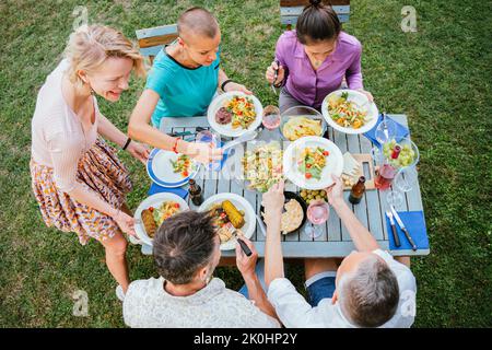 High angle view of adult friends sitting together at table, putting food on plates while having holiday dinner at backyard Stock Photo