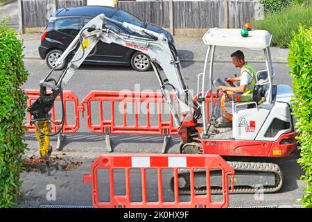 Mini digger back actor & driver breaking up pavement between saw cuts for narrow shallow fibre optic broadband trench house drive closed England UK Stock Photo