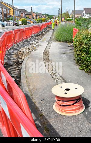 Workmen working on pavement reinstating fibre optic cable trench for new broadband infrastructure behind red interlocking safety barriers England UK Stock Photo