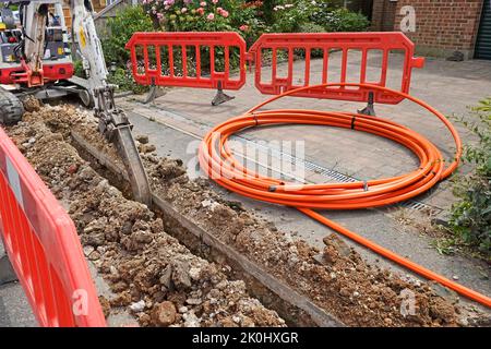 Mini digger excavator back actor machine trenching narrow channel in paving for orange fibre broadband cable coiled closed house driveway England  UK Stock Photo