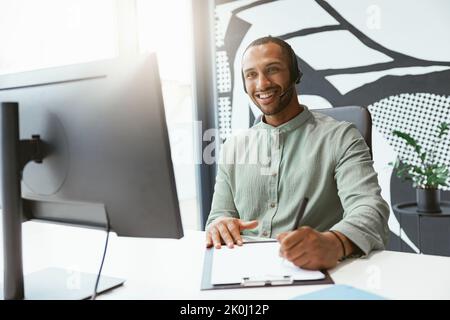 Smiling african businessman working on computer and making notes while sitting in modern office Stock Photo