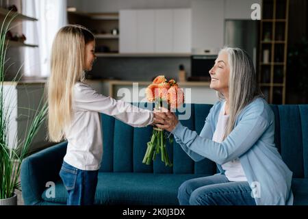 Smiling caucasian little granddaughter gives bouquet of flowers to old grandma on sofa in living room interior, profile Stock Photo