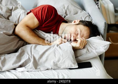 Young tired man sleeping in his bed under blanket in the bedroom Stock Photo