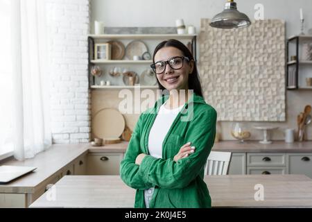 Portrait of a young beautiful brunette woman in glasses and a green shirt. Standing in the kitchen at home, looking at the camera, smiling, folded her arms over her chest. Stock Photo