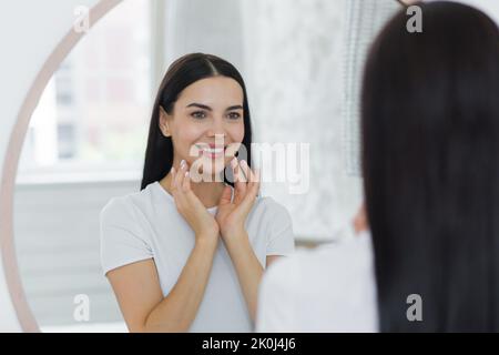 Portrait of a young beautiful brunette woman in a white T-shirt looking in the mirror at home, touching her face, skin, smiling, admiring. Stock Photo