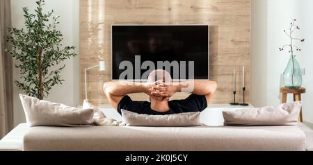 A man in a pose of reflection on the sofa in the living room in front of the TV Stock Photo