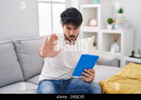 Hispanic man with beard using touchpad sitting on the sofa doing stop gesture with hands palms, angry and frustration expression Stock Photo