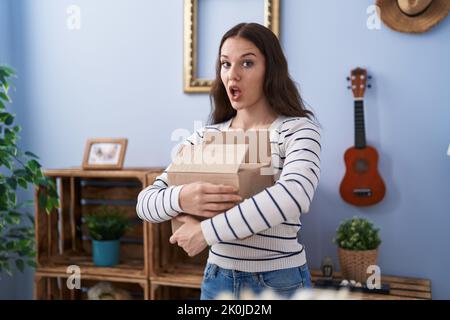 Young hispanic girl opening cardboard box in shock face, looking skeptical and sarcastic, surprised with open mouth Stock Photo