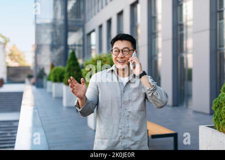Happy successful asian businessman talking on smartphone, standing outdoors against modern office center, free space Stock Photo