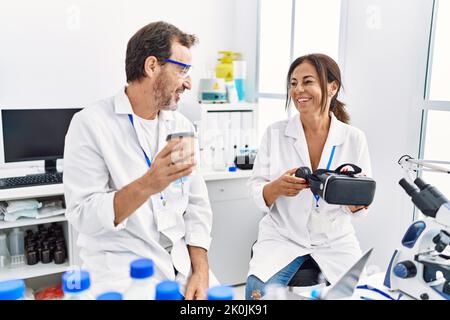 Middle age man and woman partners wearing scientist uniform drinking coffee and holding vr goggles at laboratory Stock Photo