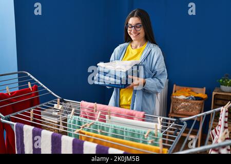 Young hispanic woman holding folded jeans hanging clothes on clothesline at laundry room Stock Photo