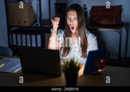 Young brunette woman working at the office at night angry and mad raising fist frustrated and furious while shouting with anger. rage and aggressive c Stock Photo