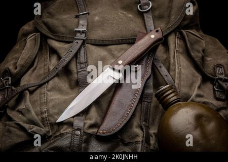 A hunting knife with a wooden handle and a leather sheath on a khaki canvas backpack. Weapons for self-defense and survival. Dark background. Stock Photo