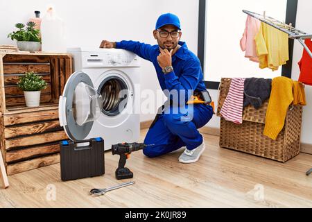 Young indian technician working on washing machine looking confident at the camera smiling with crossed arms and hand raised on chin. thinking positiv Stock Photo