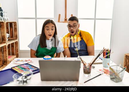 Young hispanic couple using laptop sitting on the table at art studio scared and amazed with open mouth for surprise, disbelief face Stock Photo