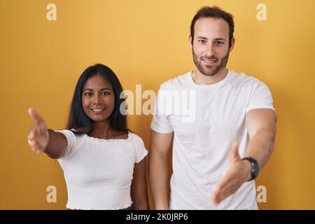 Interracial couple standing over yellow background smiling friendly offering handshake as greeting and welcoming. successful business. Stock Photo
