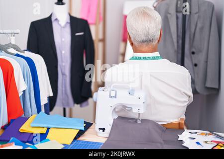 Middle age man with grey hair dressmaker using sewing machine standing backwards looking away with crossed arms Stock Photo
