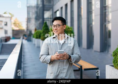 Positive asian middle aged male entrepreneur checking emails on smartphone while walking by business center Stock Photo