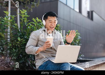 Excited asian middle aged businessman working on laptop outdoors, wearing headphones and having video call Stock Photo