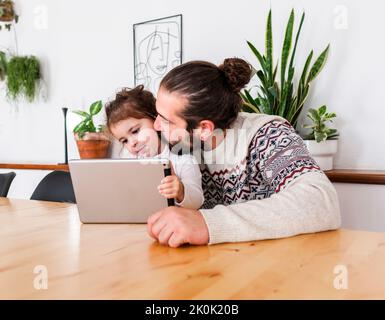 Bearded male in sweater sitting at table and kissing on cheek daughter using tablet in light apartment with potted plants Stock Photo