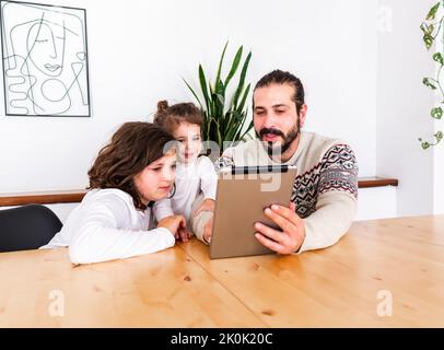 Happy father with beard in warm sweater sitting at table with children while using tablet in apartment Stock Photo