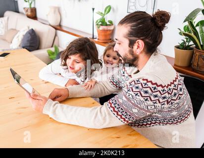 Happy father with beard in warm sweater sitting at table with children while using tablet in apartment Stock Photo